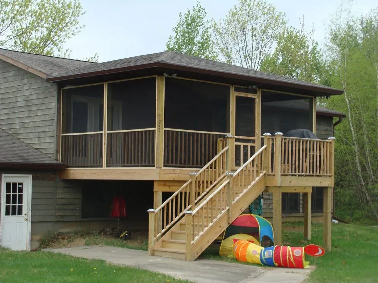 Wooden screened porch attached to a house with stairs leading to the ground, with playground equipment below.