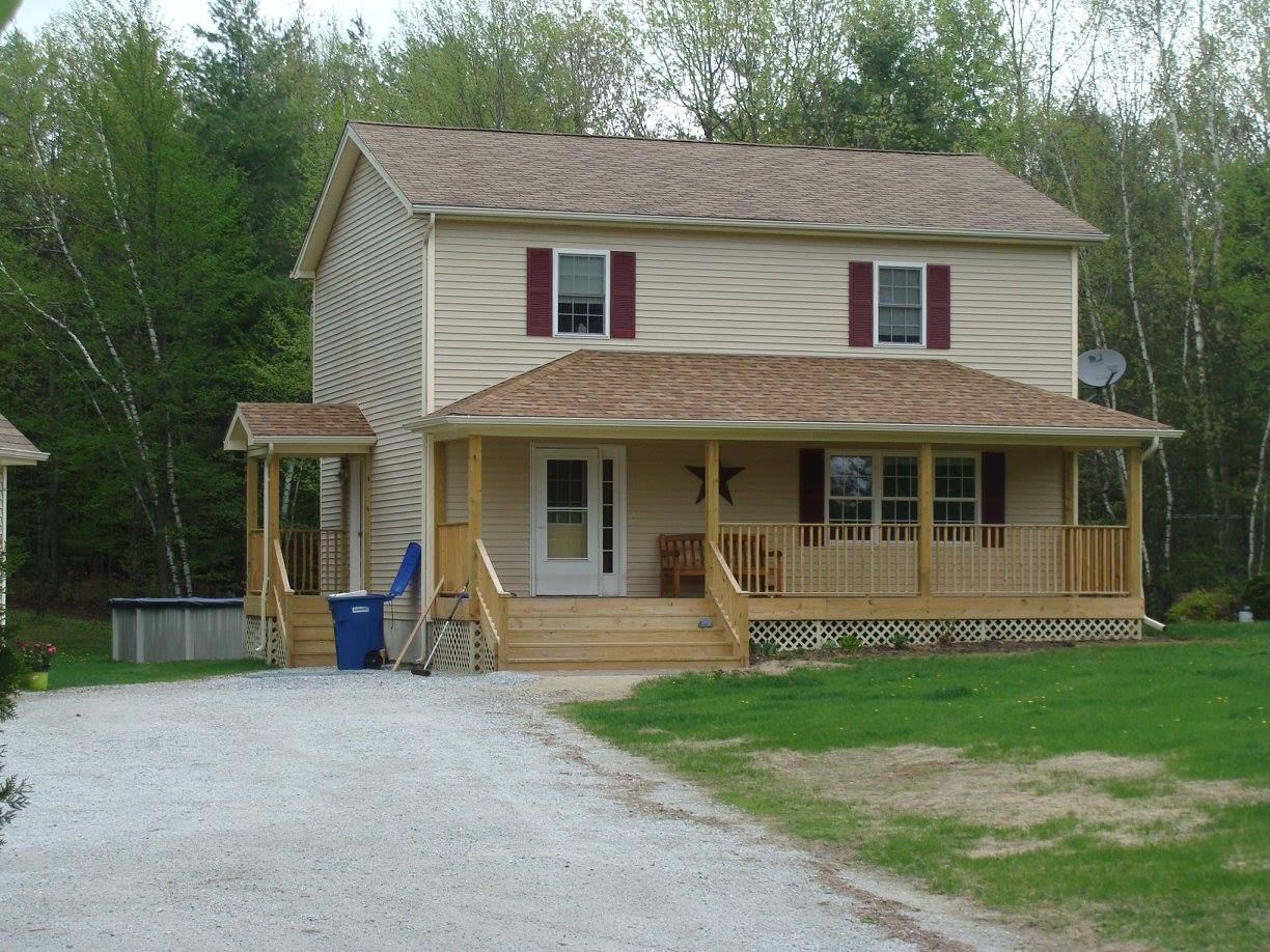 Two-story beige house with red shutters and a wooden porch, gravel driveway, and green lawn.