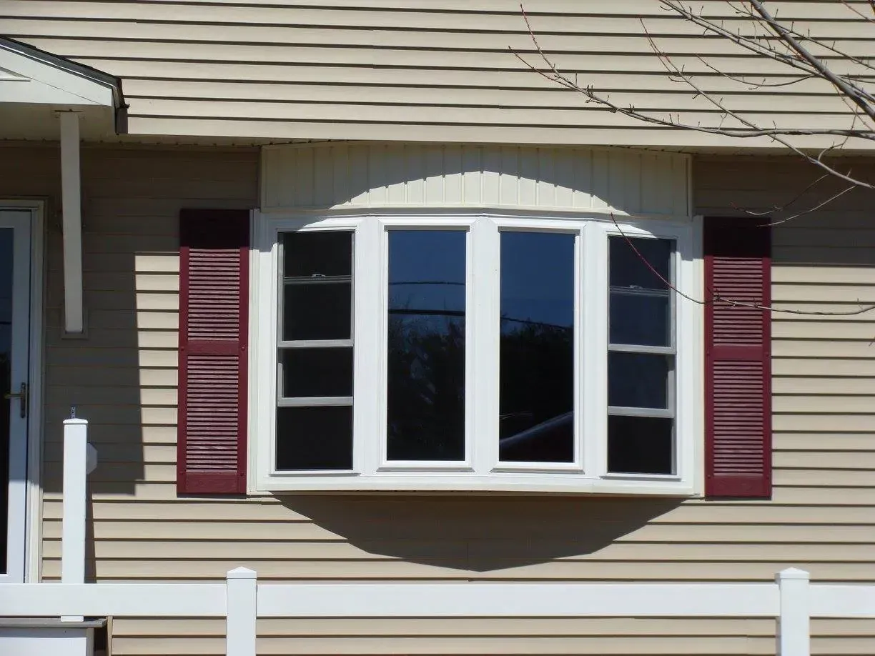 A bay window with red shutters on a beige house, with white trim and a shadow on the wall.