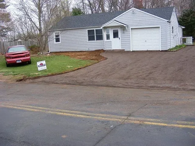 Ranch-style house with gray siding and a newly laid driveway, a red car parked nearby.