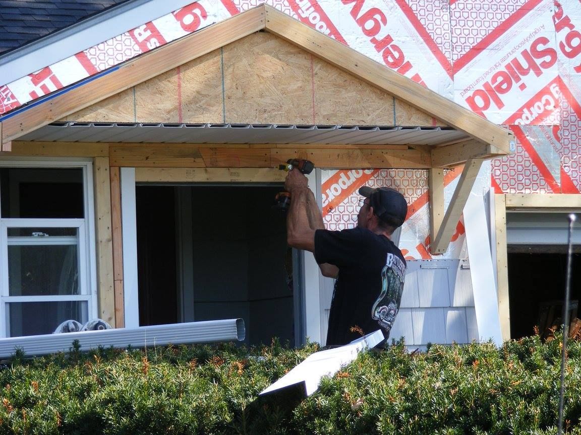 Construction worker hammering a wood beam on a house, under a partially built porch. Red siding visible.