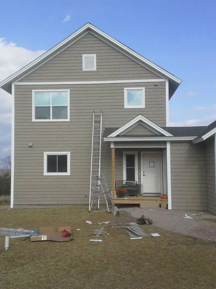 Two-story house with ladder propped against it; gray siding, white trim, door under a small porch, supplies on the ground.