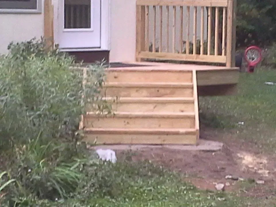 Wooden steps leading up to a deck and door. Green foliage surrounds the steps.