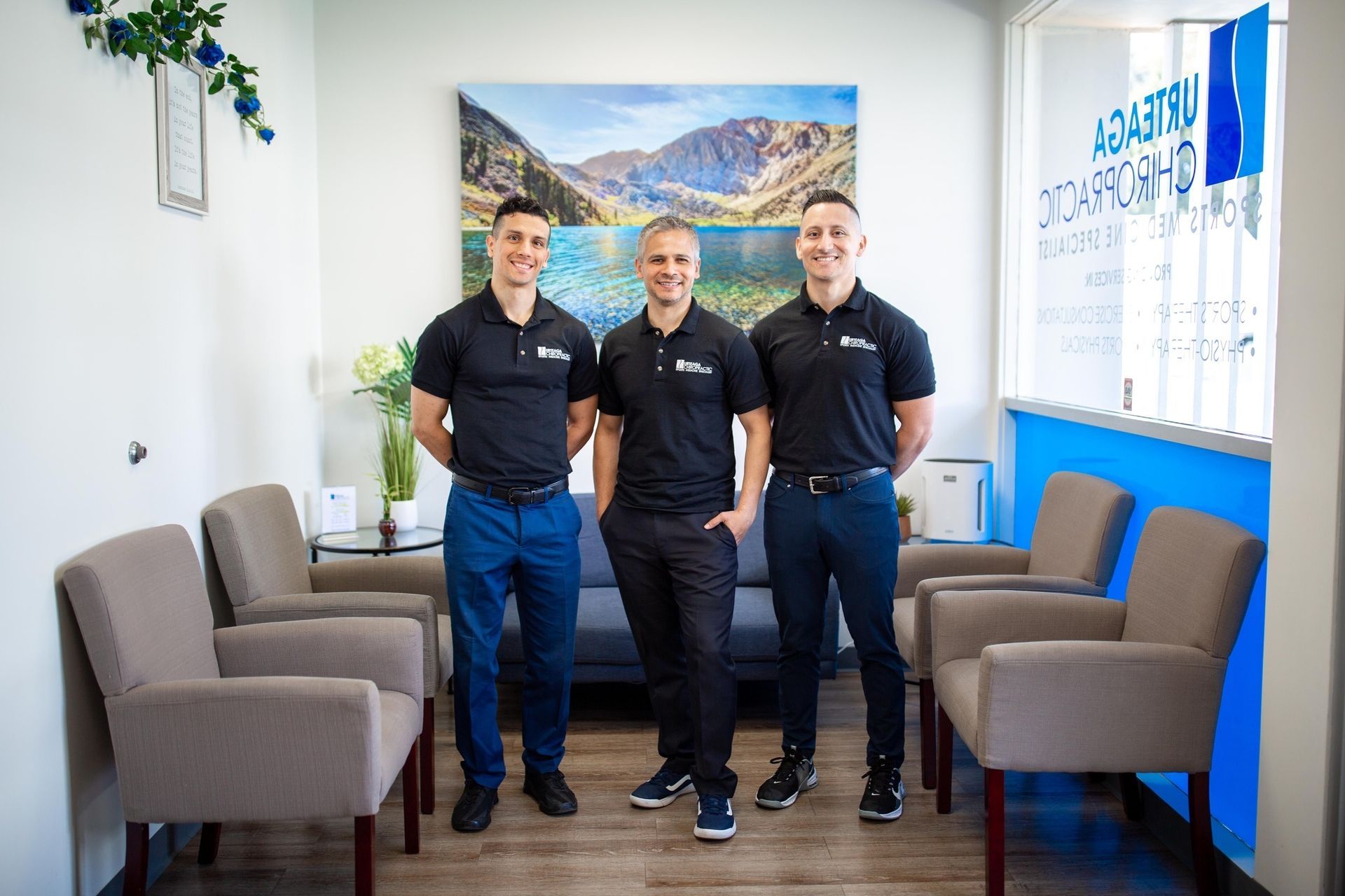 Three men in black shirts stand in a waiting room. Mountain painting, blue and gray chairs.
