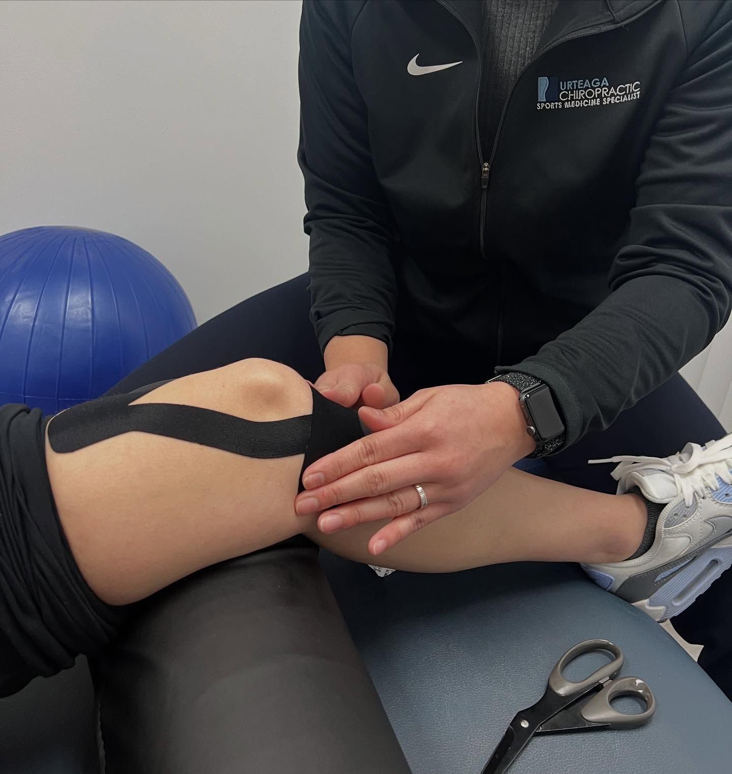 Person applying black kinesiology tape to a knee, indoors. Scissors nearby.