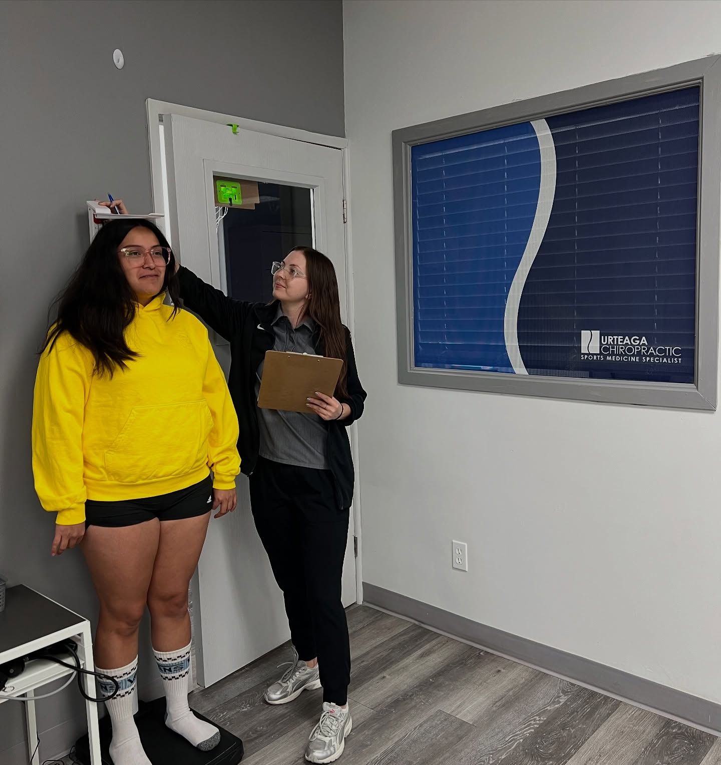 Woman in yellow hoodie being measured by another woman in an office; grayscale scale, white door, blue and white window.