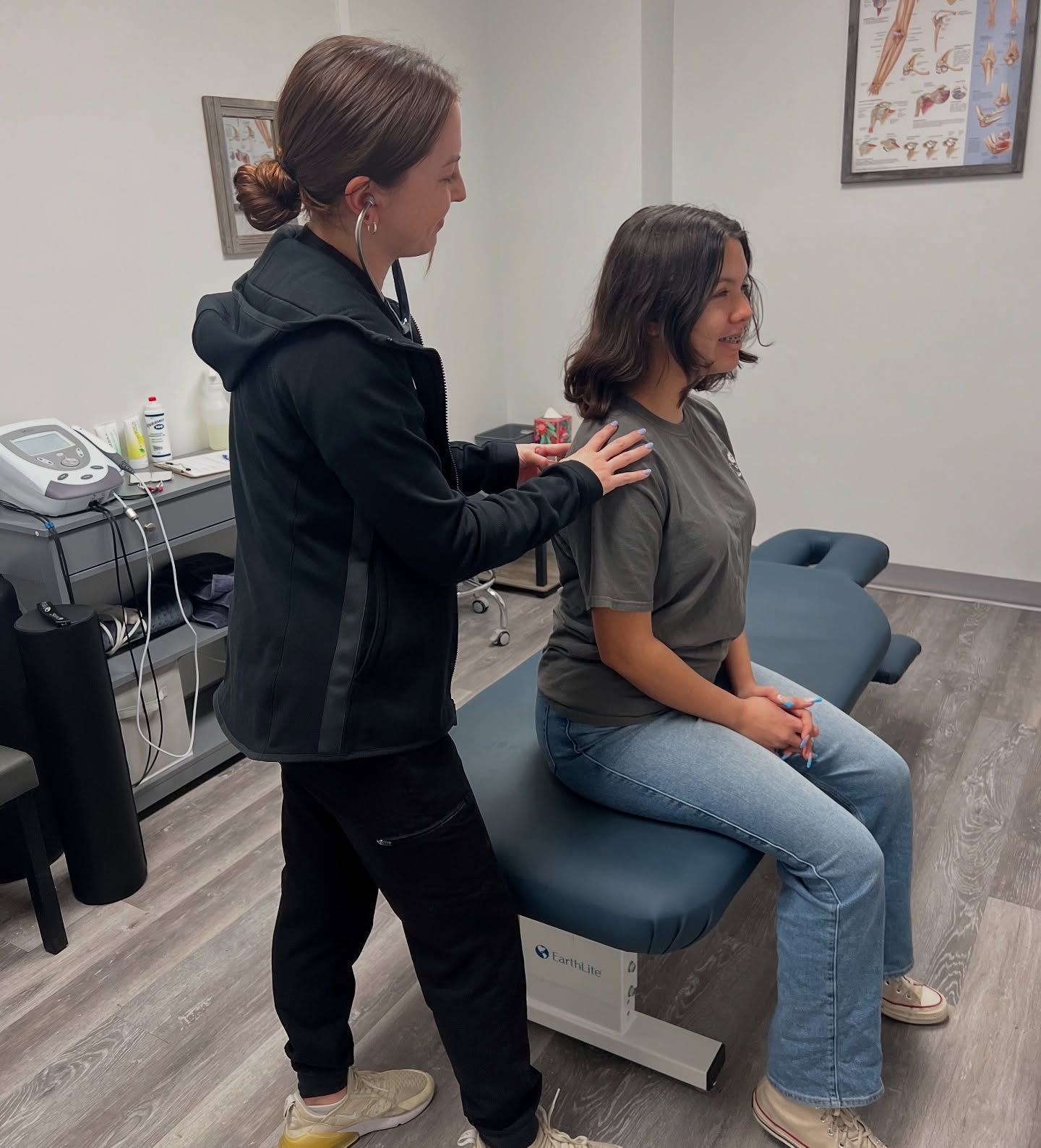 A healthcare professional assesses a seated patient's shoulders. Indoors, neutral tones.