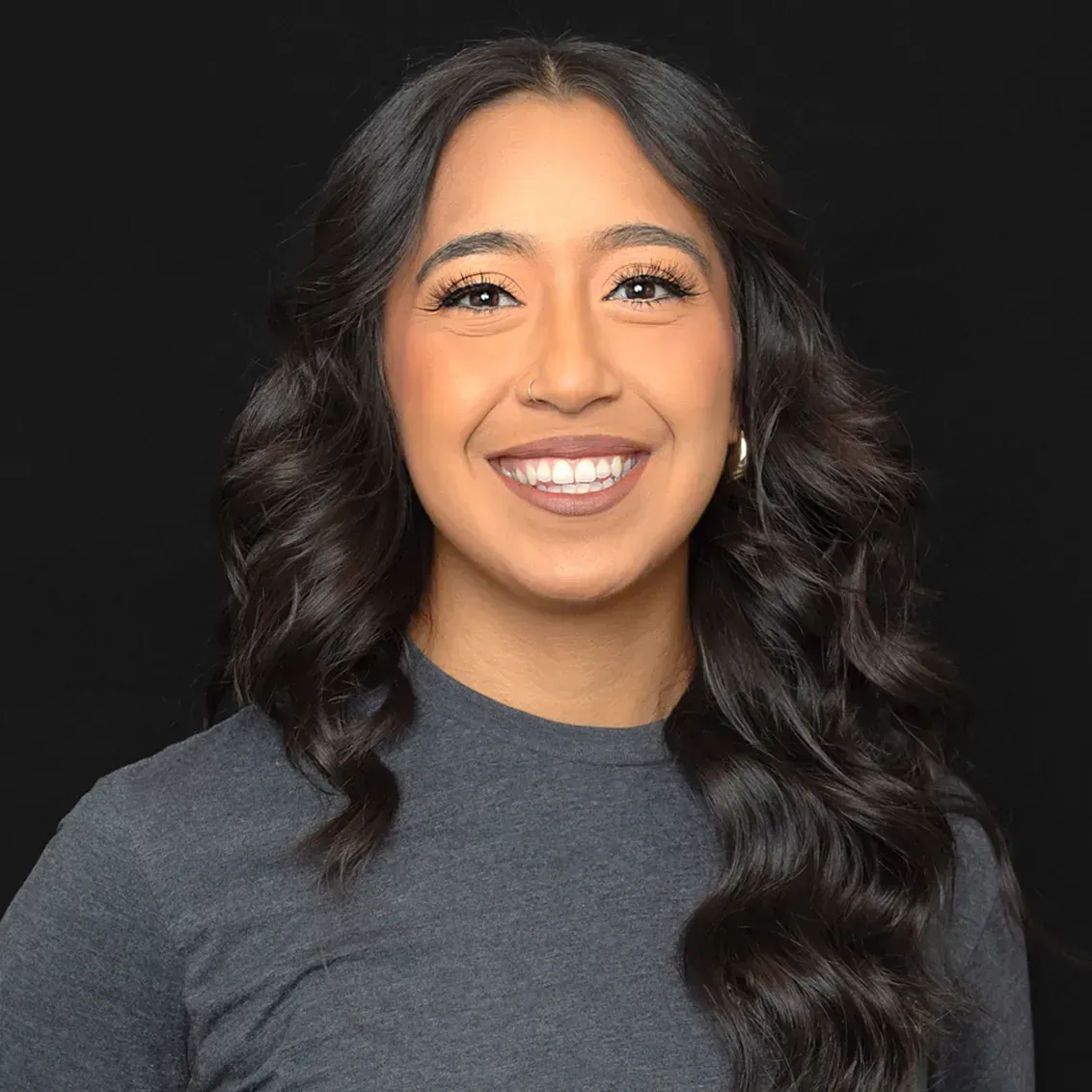 Smiling woman with dark hair, wearing a gray shirt, against a black backdrop.