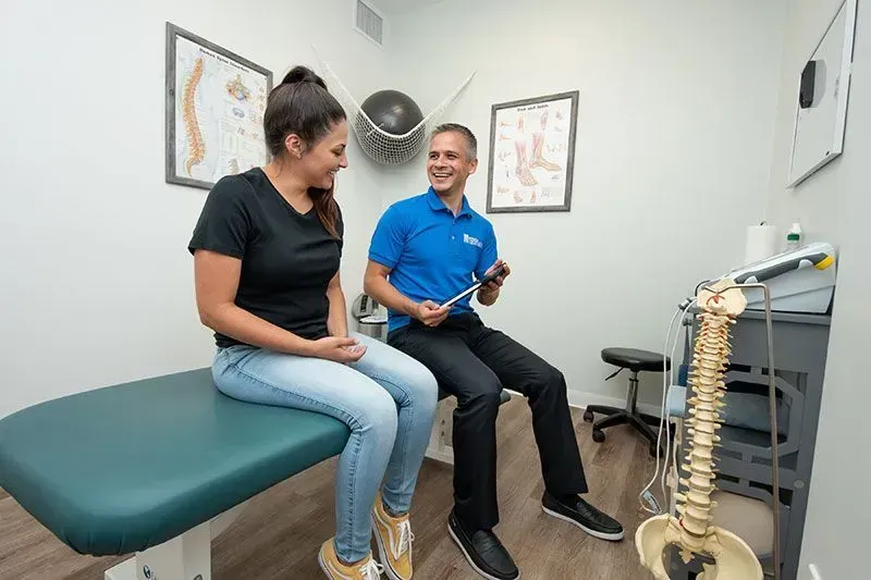 A doctor examines a patient's back in an office. Both are smiling, sitting. A spine model is next to the equipment.