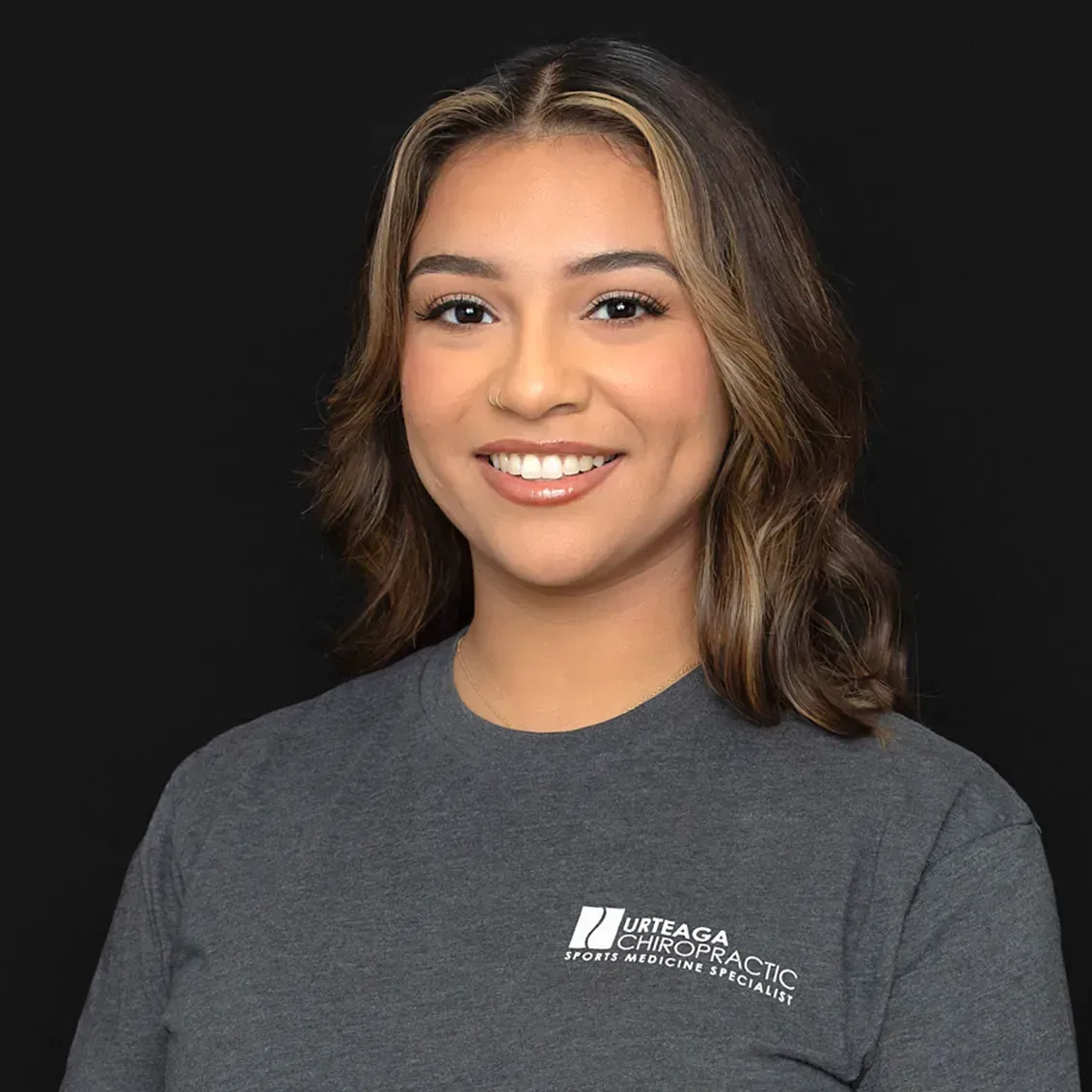 Woman with brown hair wearing gray shirt smiles at the camera. The background is black.