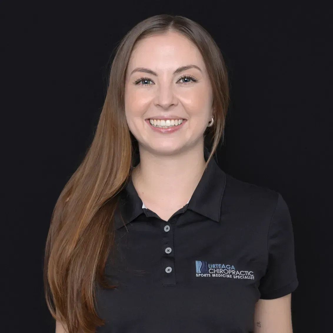Woman smiling, wearing a black polo shirt with a logo. Long brown hair, black background.