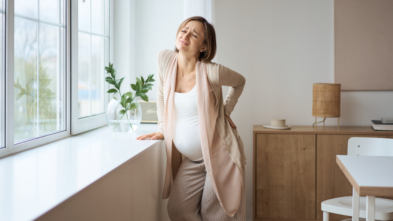 Pregnant woman leans against a window sill, clutching her back in pain. White tank top, beige sweater.