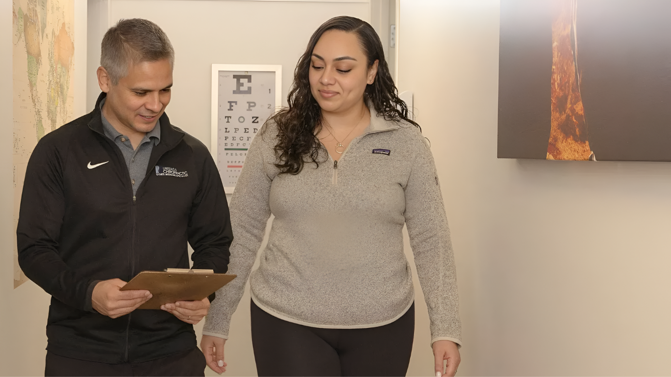 A man in black jacket and woman in gray sweater walk in a medical office, man holding a clipboard.