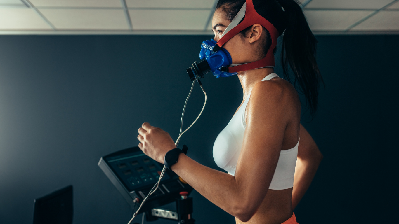 Woman runs on a treadmill wearing a mask and connected to equipment for testing; white top, dark hair.
