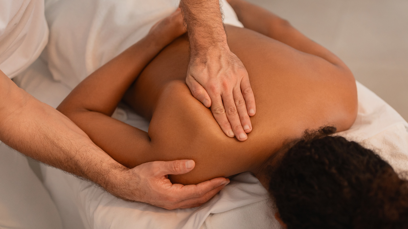 Person receiving a shoulder massage from a masseuse, on a white sheet.