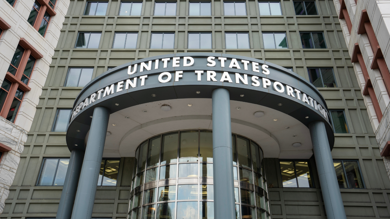 United States Department of Transportation building entrance with sign above revolving doors. Gray facade, cloudy sky.