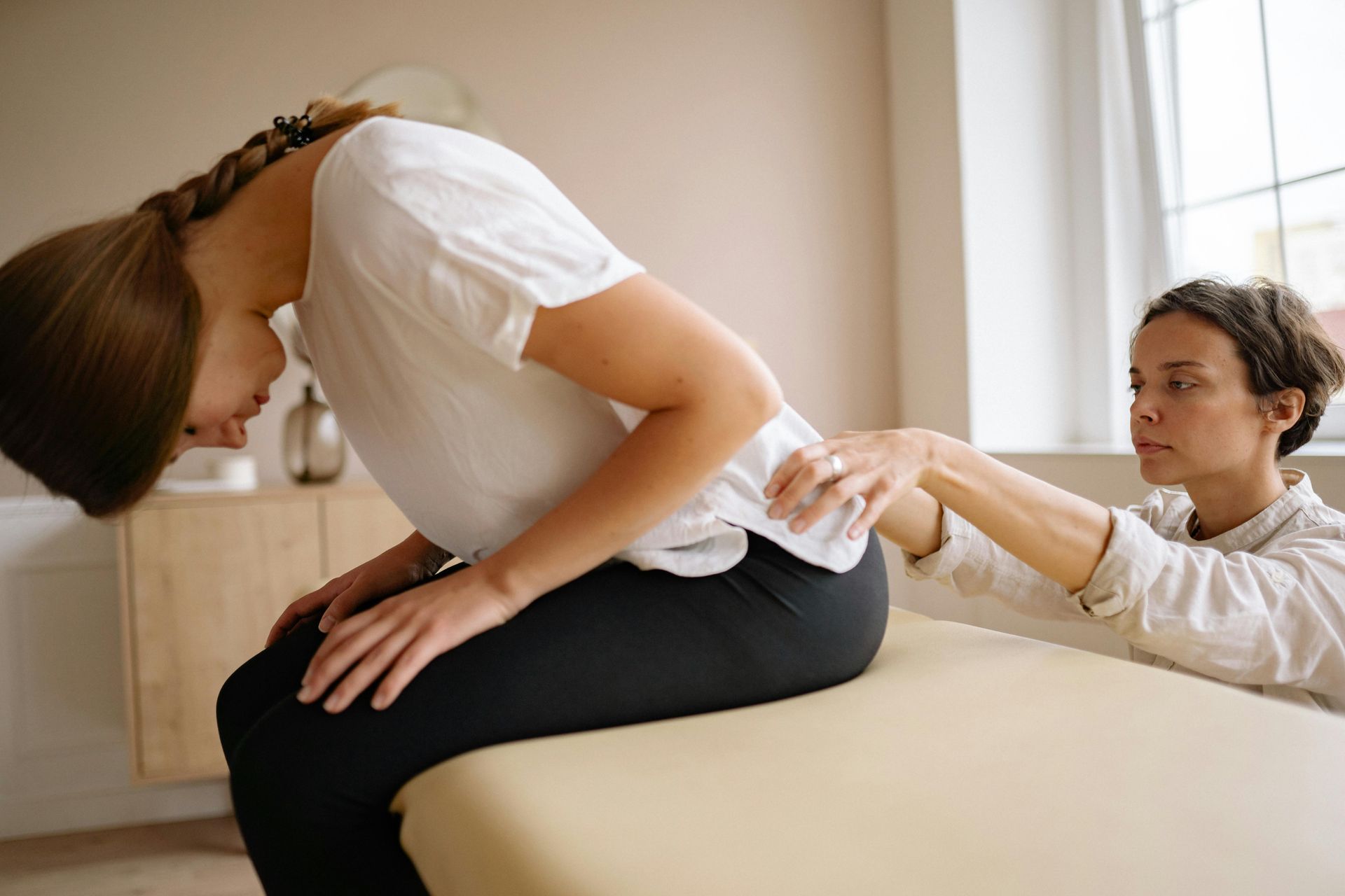 Woman in white shirt and black pants receiving a seated back stretch from a therapist in a bright room