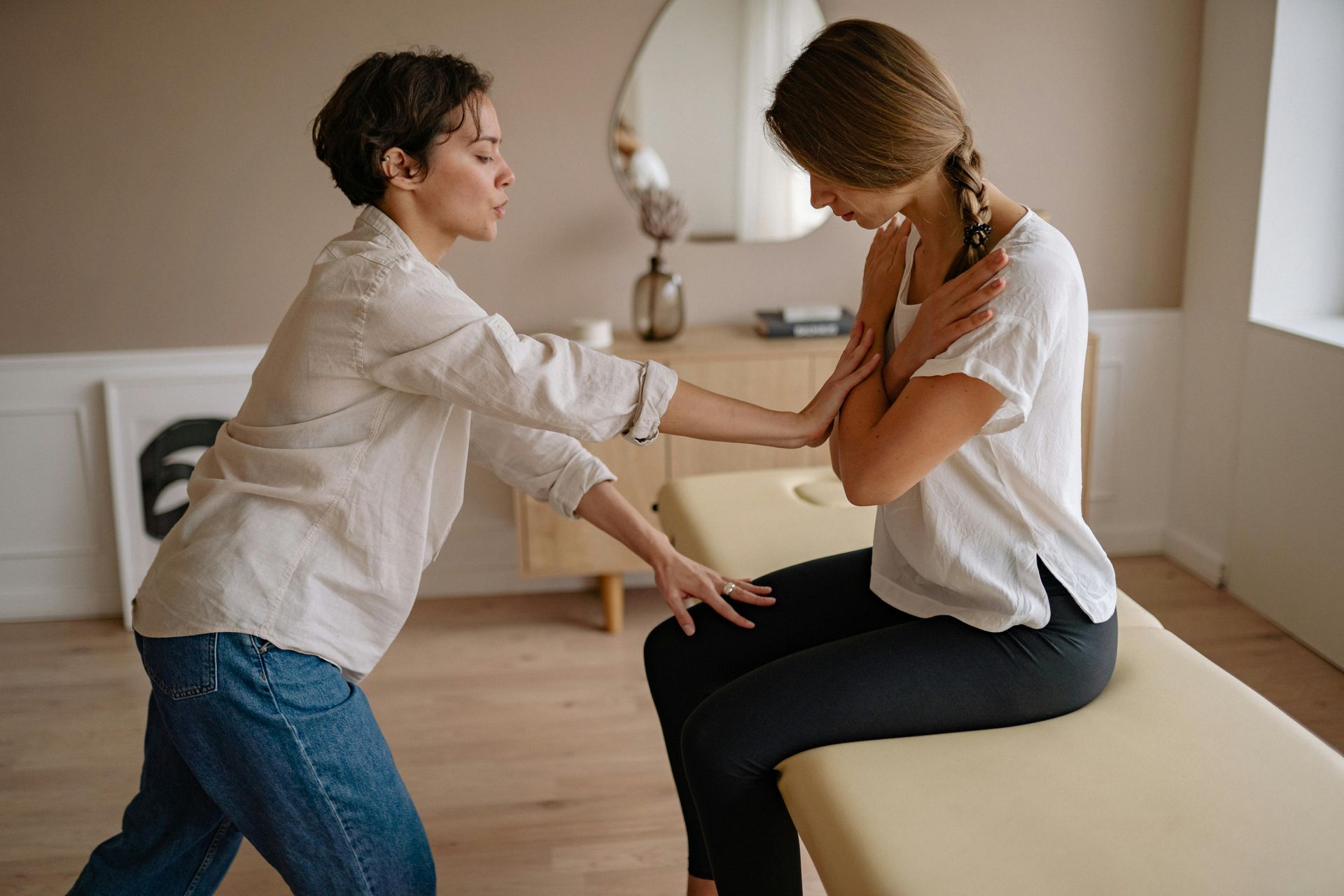 Woman examines another woman's arm while she's sitting on a massage table in a room.