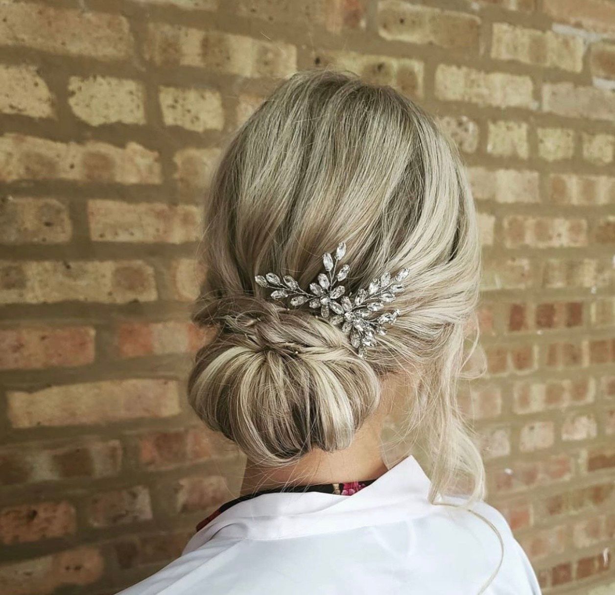 A woman with blonde hair in a bun with a comb in her hair is standing in front of a brick wall.