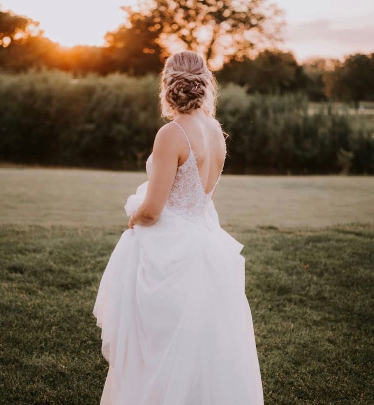 A bride in a white dress is standing in a field at sunset.