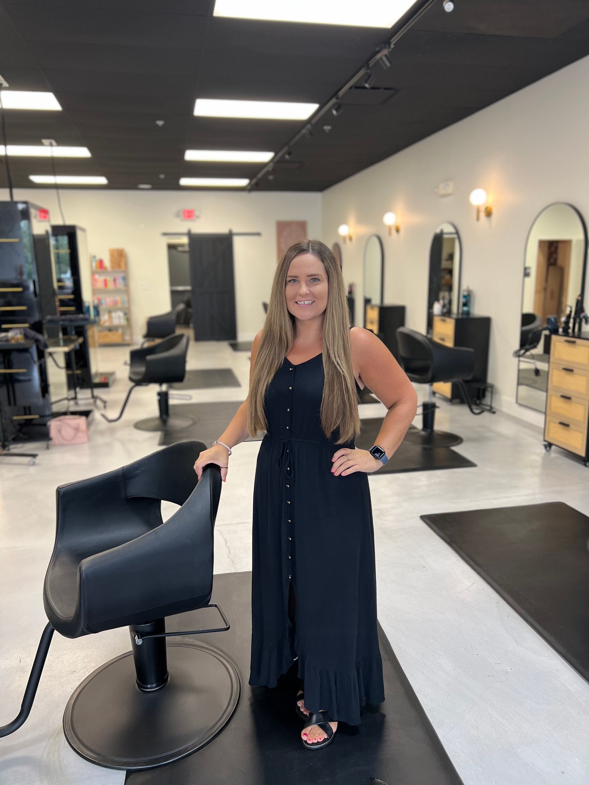 A woman in a black dress is standing next to a black chair in a salon.