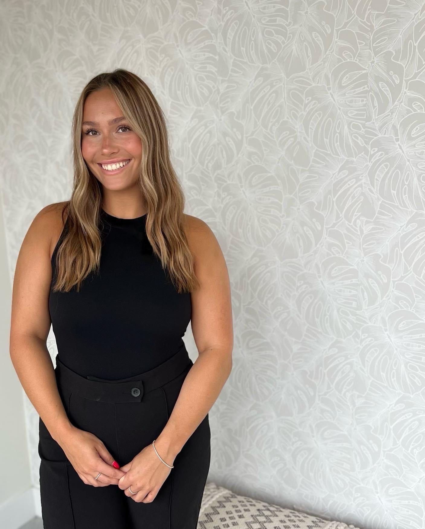 A woman in a black tank top and black pants is standing in front of a wall.
