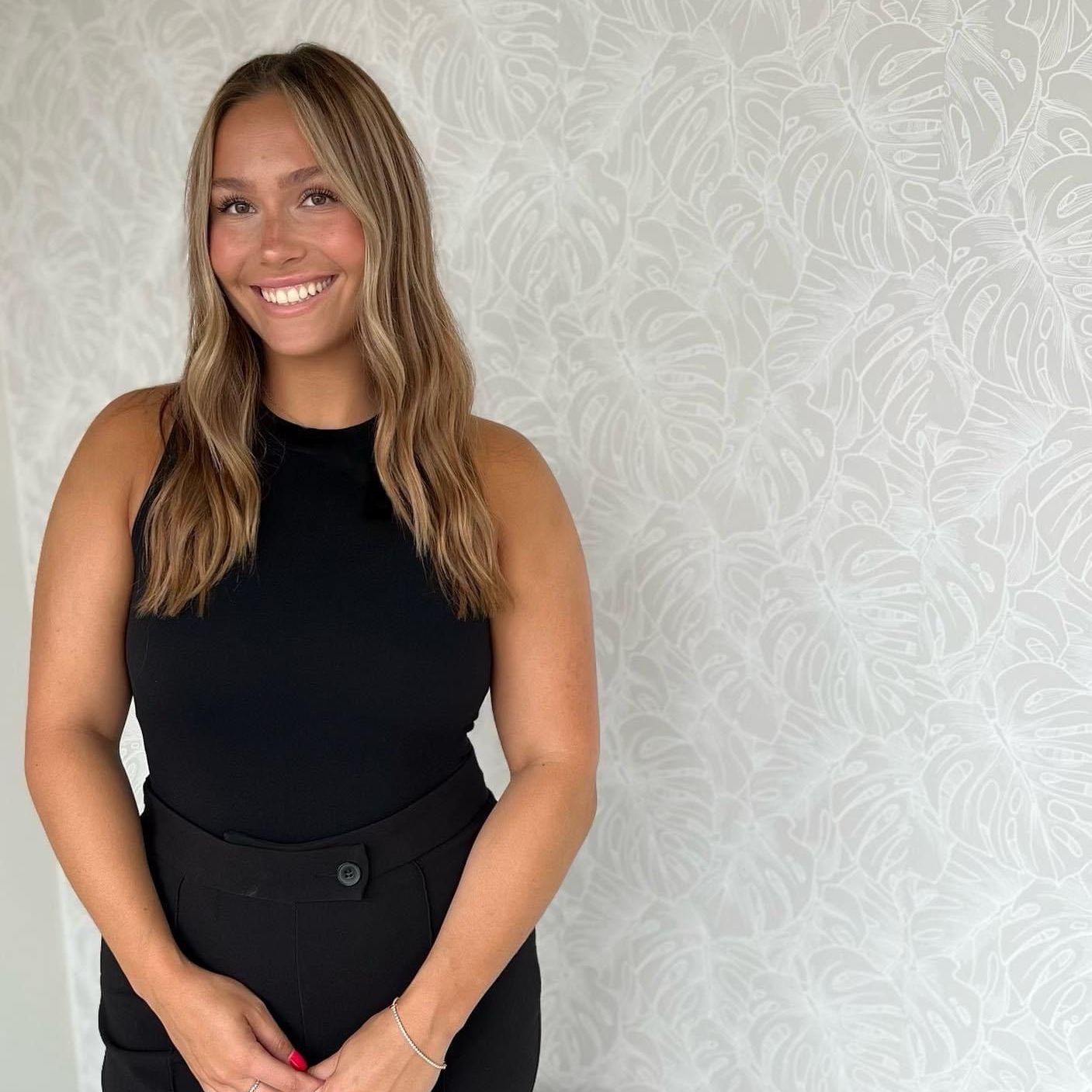 A woman in a black tank top is smiling in front of a white wall.