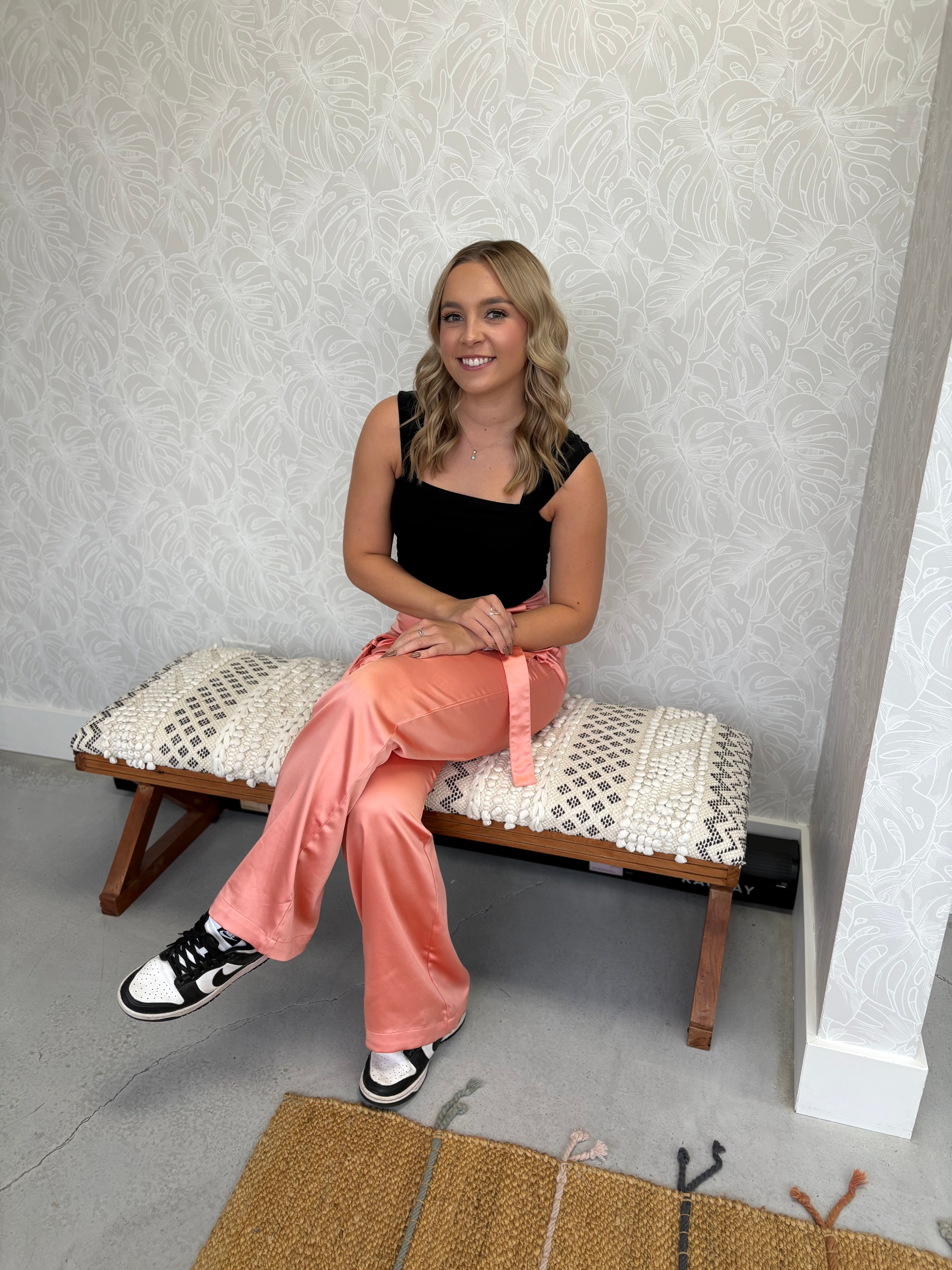 A woman in a black top and pink pants is sitting on a bench.