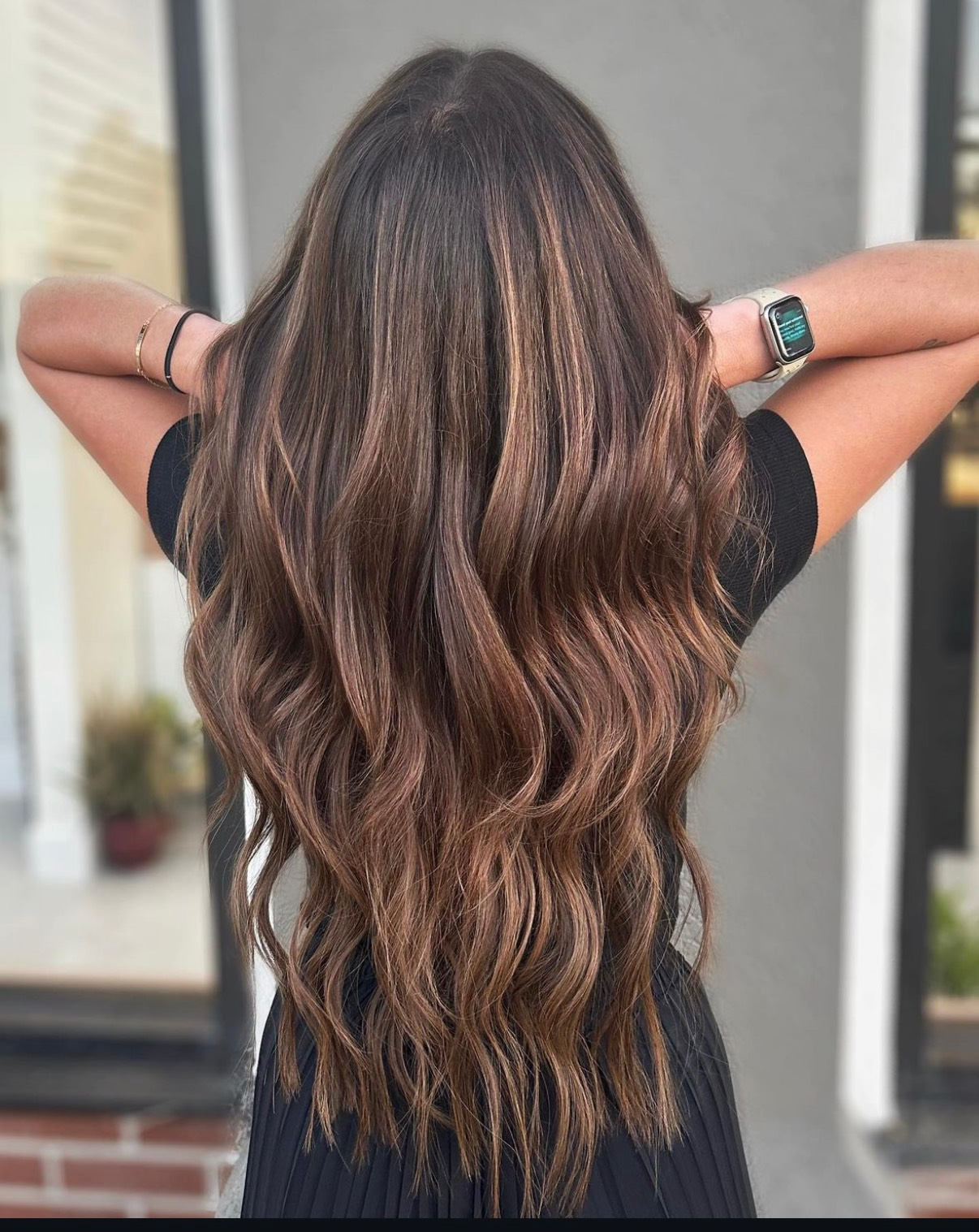 A woman with long brown hair is wearing a black dress and a watch. Customized Lived in balayage
