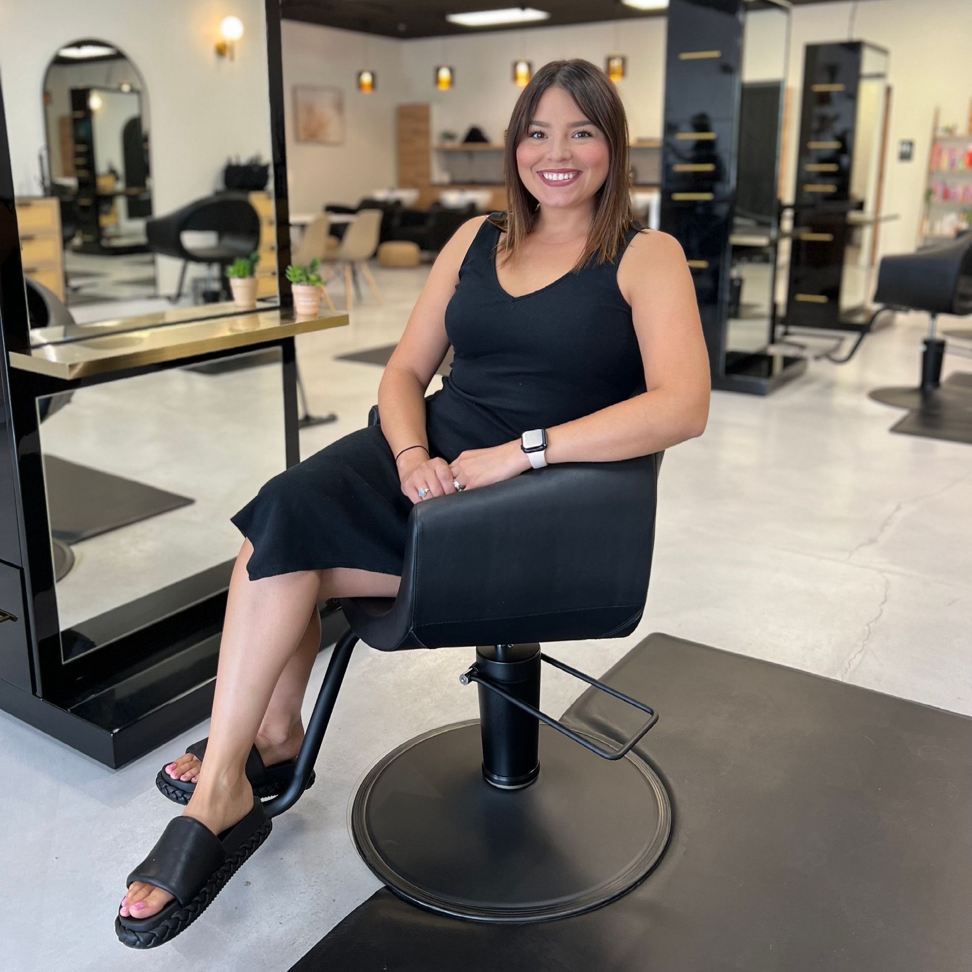 A woman in a black dress is sitting in a black chair in a salon.