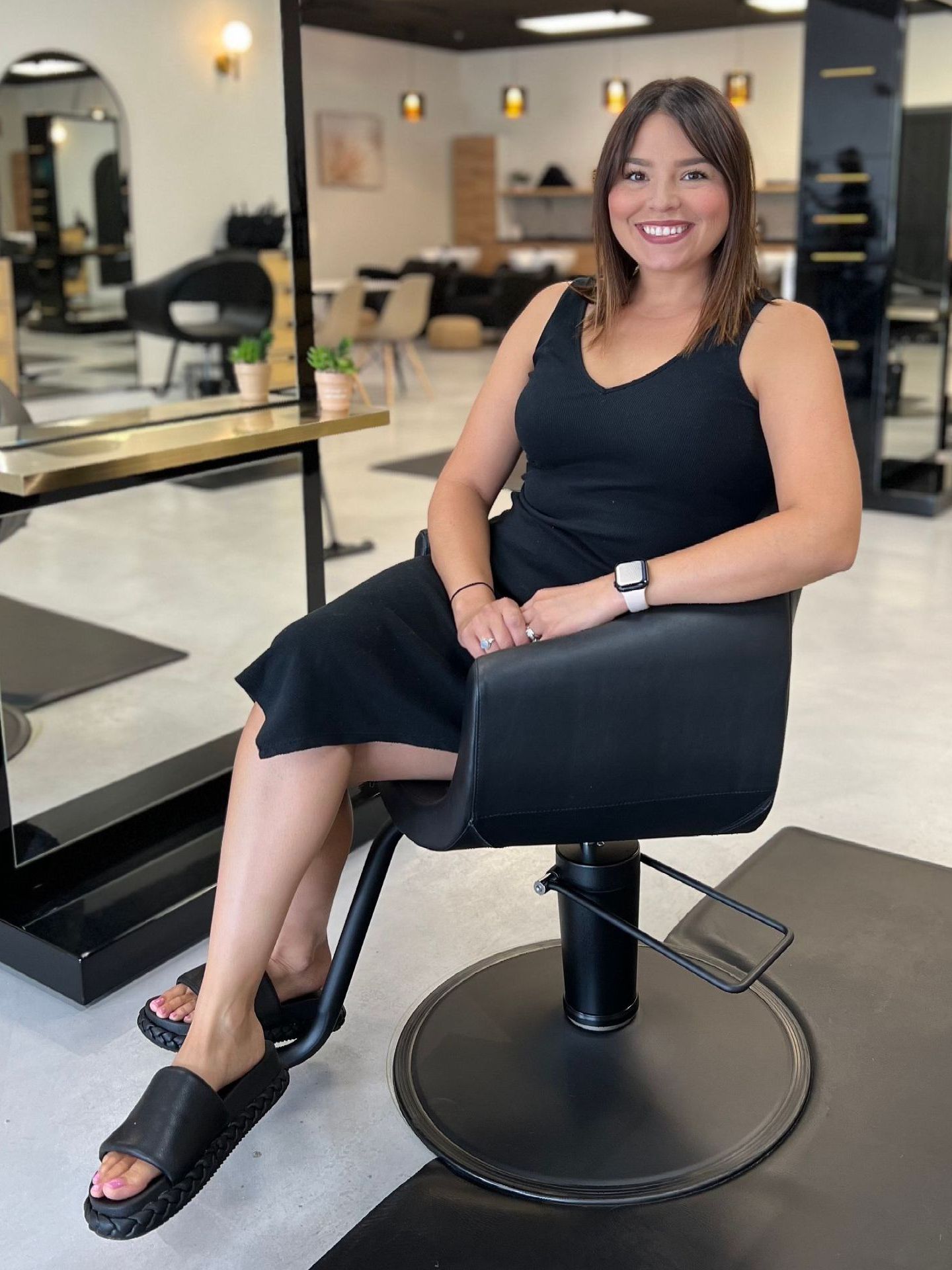 A woman in a black dress is sitting in a black chair in a salon.