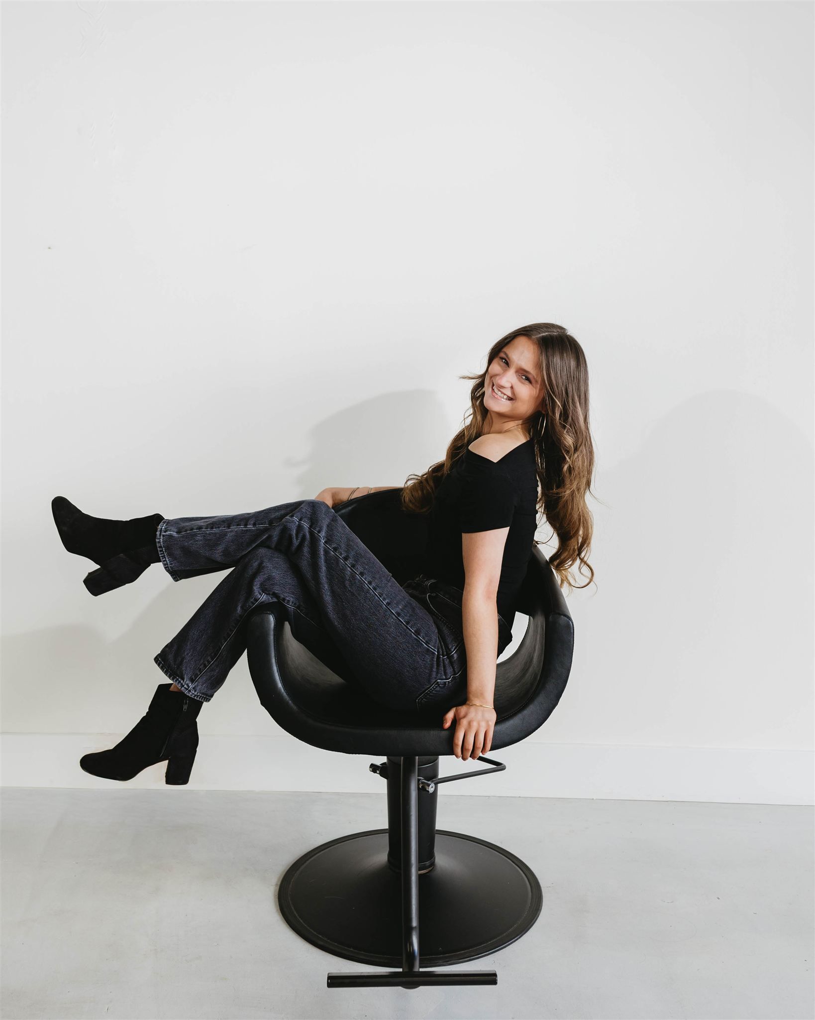 Woman smiling, sitting in black chair, legs crossed, wearing black shirt and jeans. White wall background.
