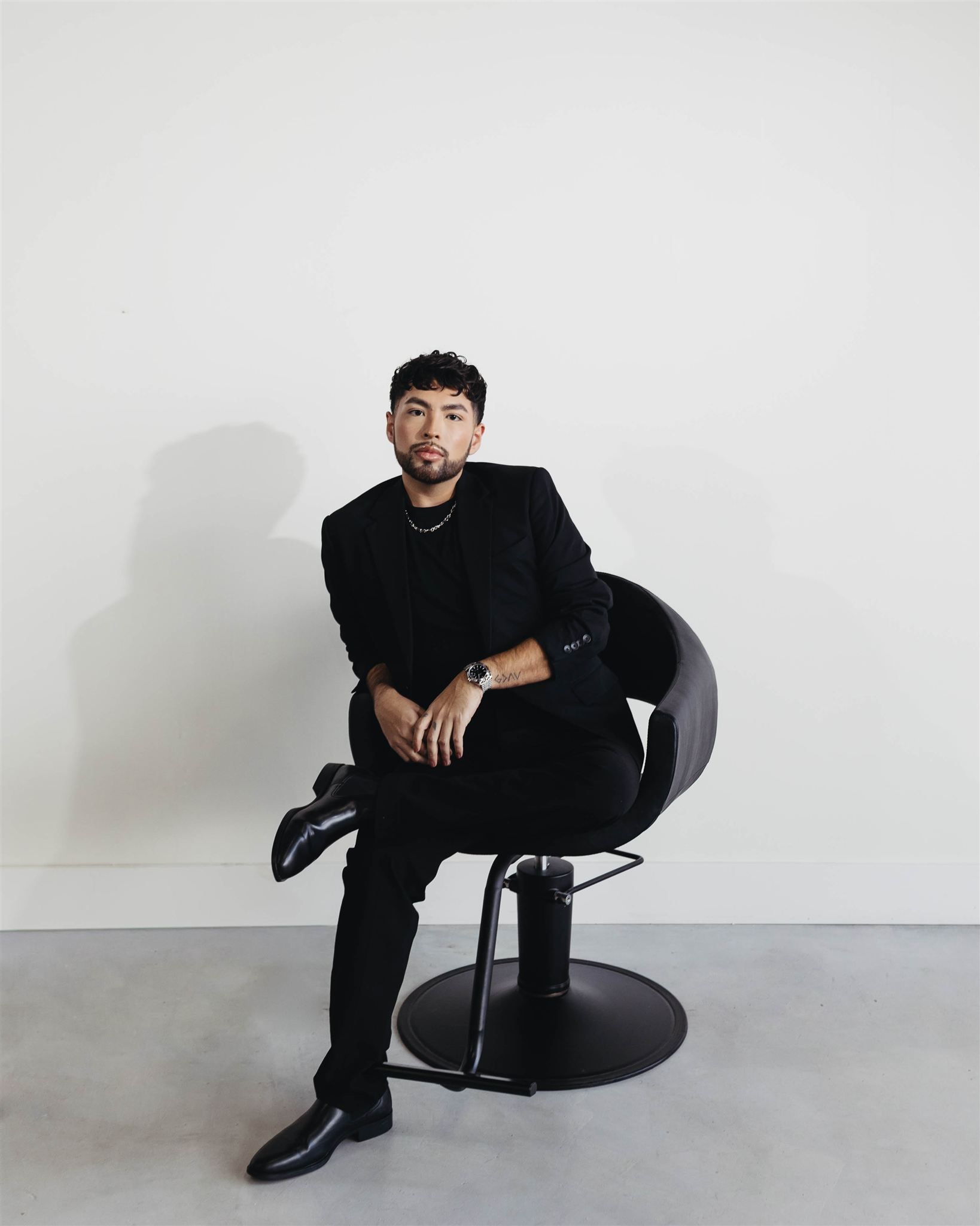 Man in black suit sits on a chair, legs crossed, against a white wall.