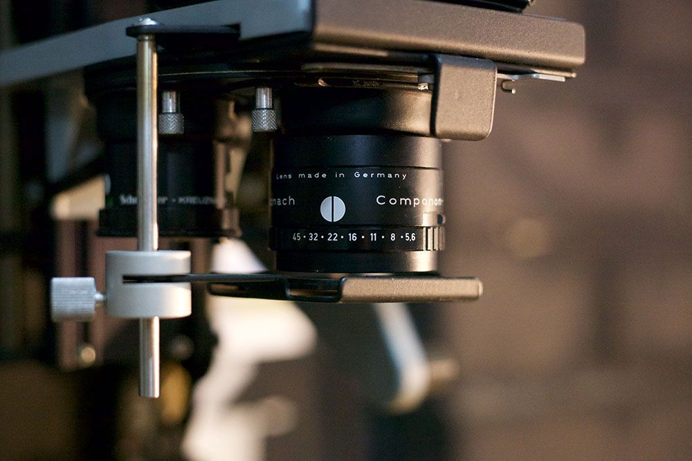 Close-up of a black enlarger lens, mounted on a metal arm.