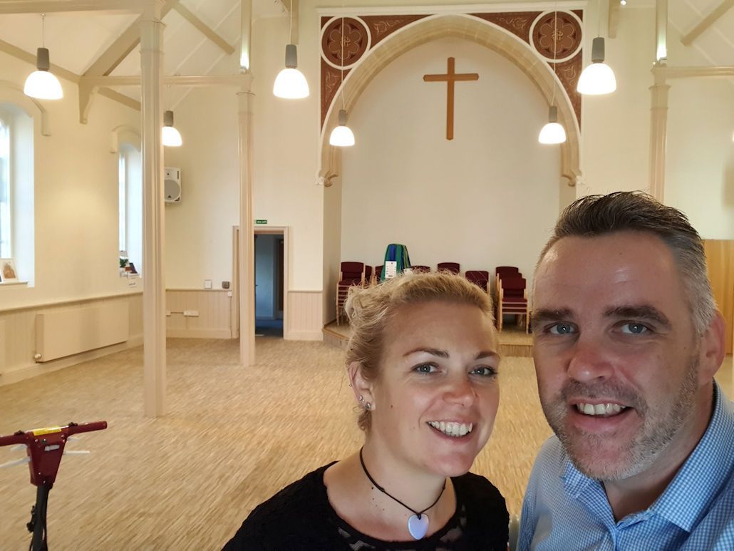 A man and a woman are posing for a picture in an empty church with a cross on the wall.