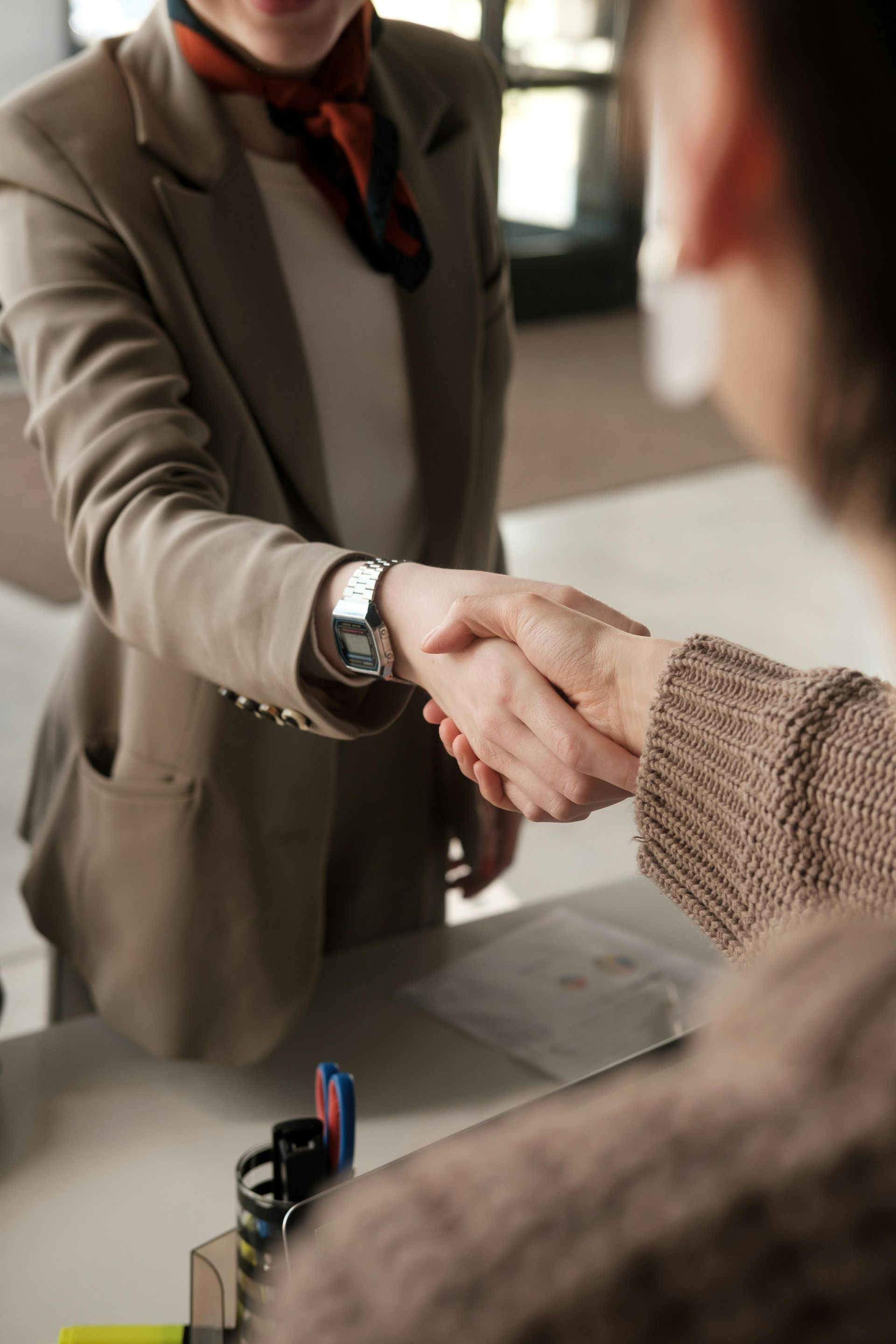 Two people shaking hands at a counter. One wears a suit with a scarf, the other a sweater.