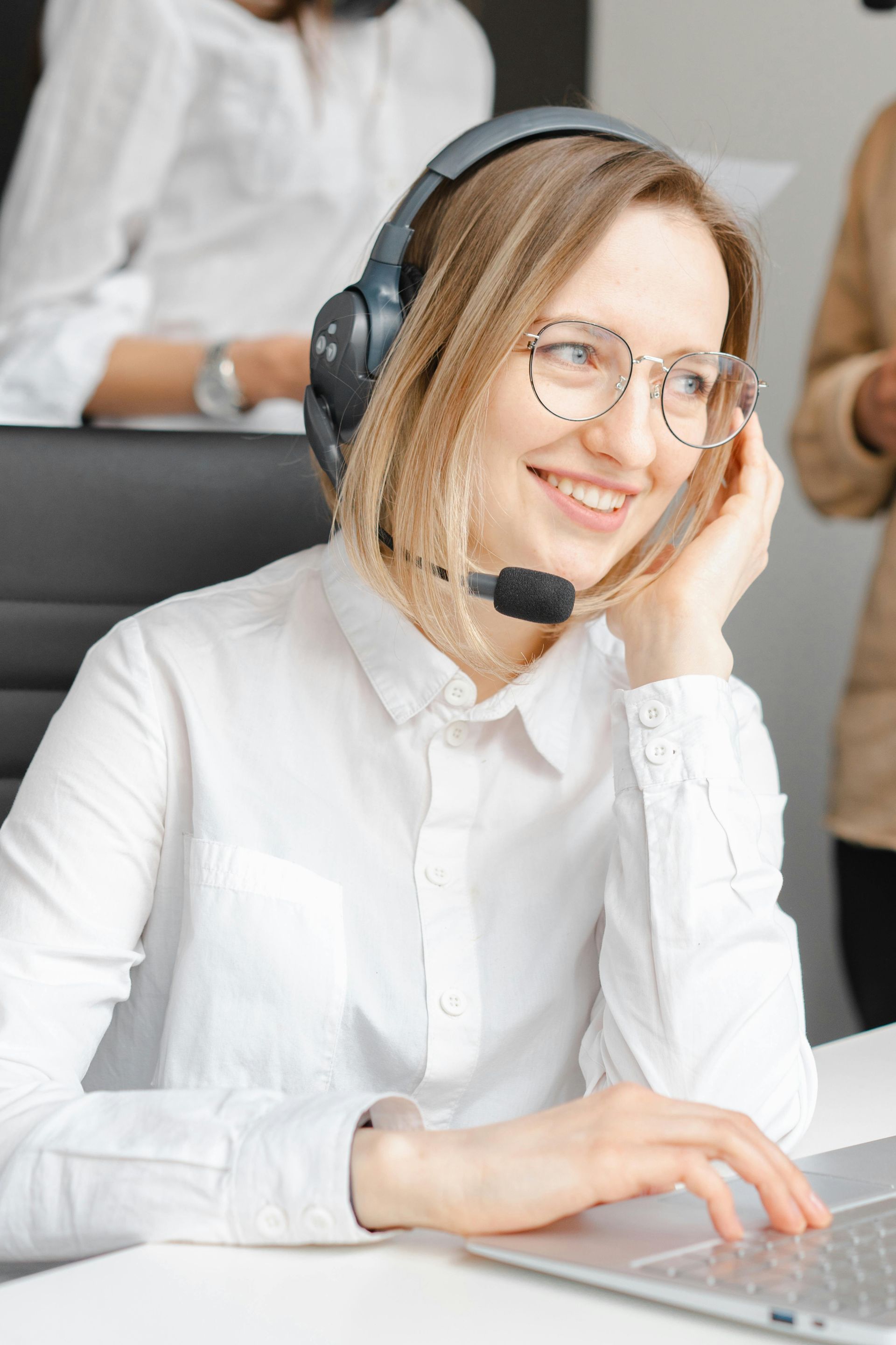 Woman with headset smiling, wearing glasses, at a desk with laptop.