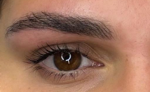 Primo piano di un occhio marrone e di un sopracciglio folto e scuro. L'occhio presenta mascara e un riflesso luminoso.