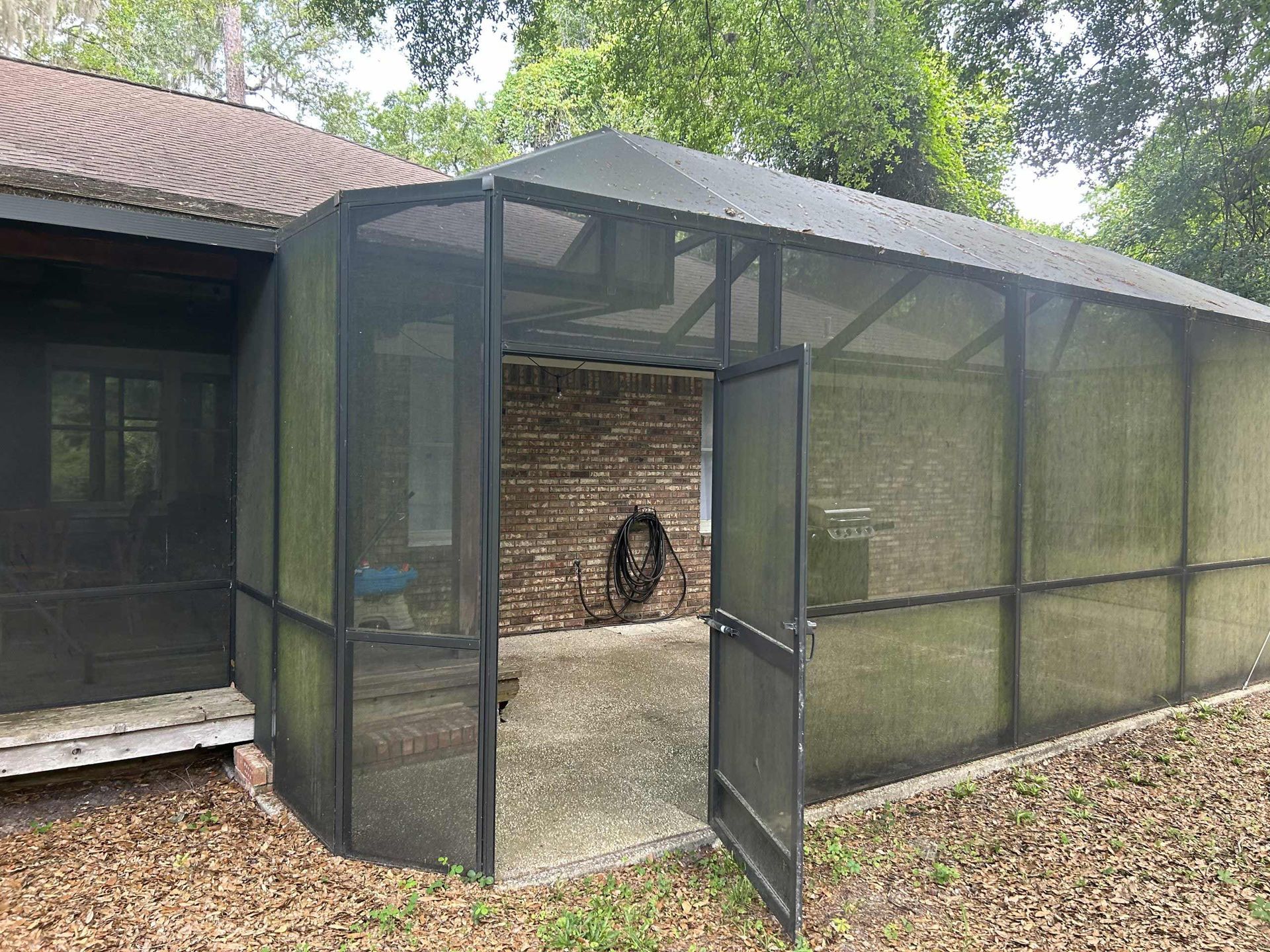 Screened porch with open door next to a brick wall. Black framing, beige concrete, and brown leaves.