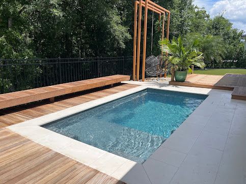 Small rectangular pool with wooden deck and bench, surrounded by a black fence and trees.