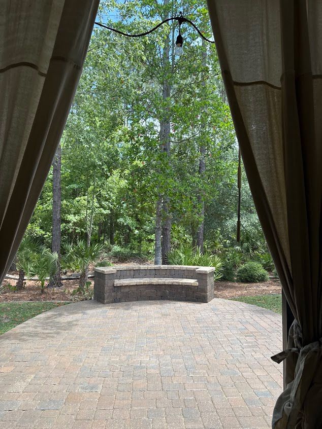 Brick patio with stone bench framed by curtains, overlooking trees.