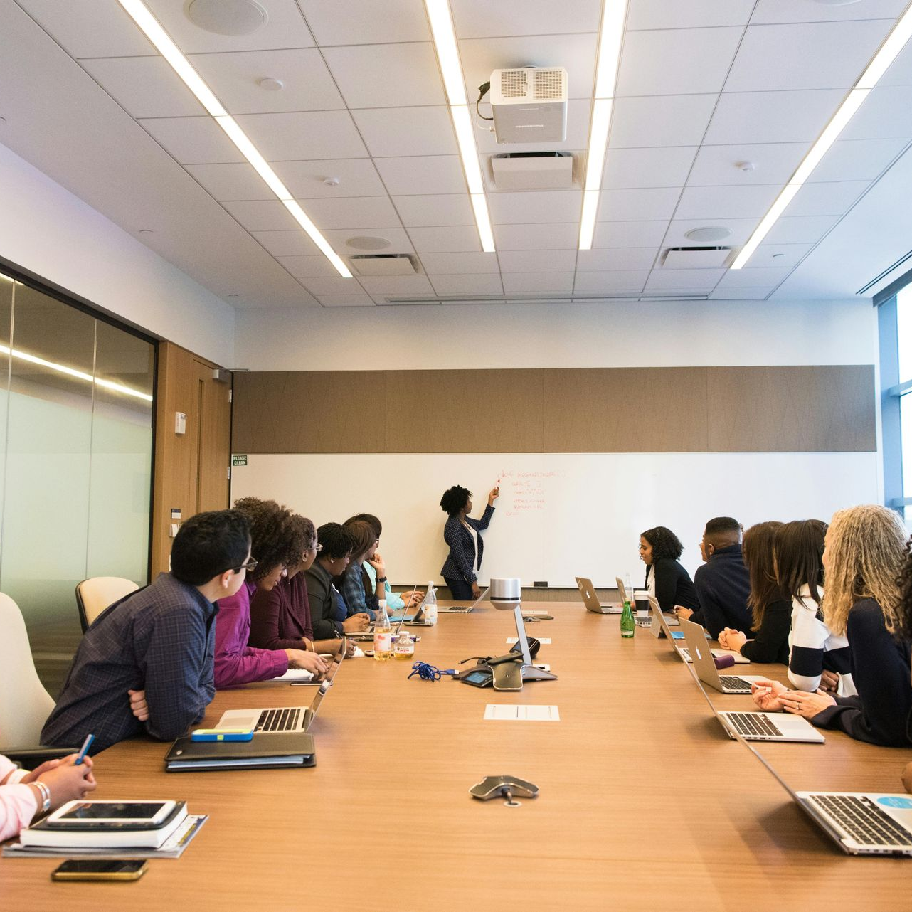 People in a boardroom. A woman writes on a whiteboard as others watch and use laptops at a large table.