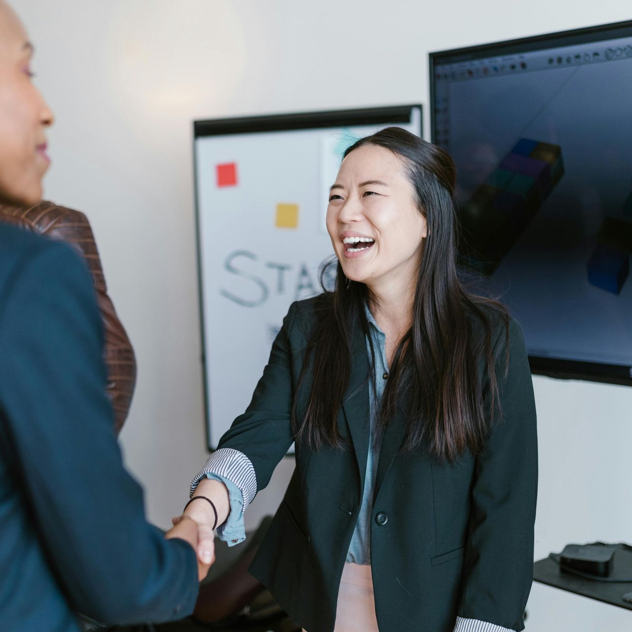 Woman shaking hands with another, smiling in an office setting with a whiteboard and screen.