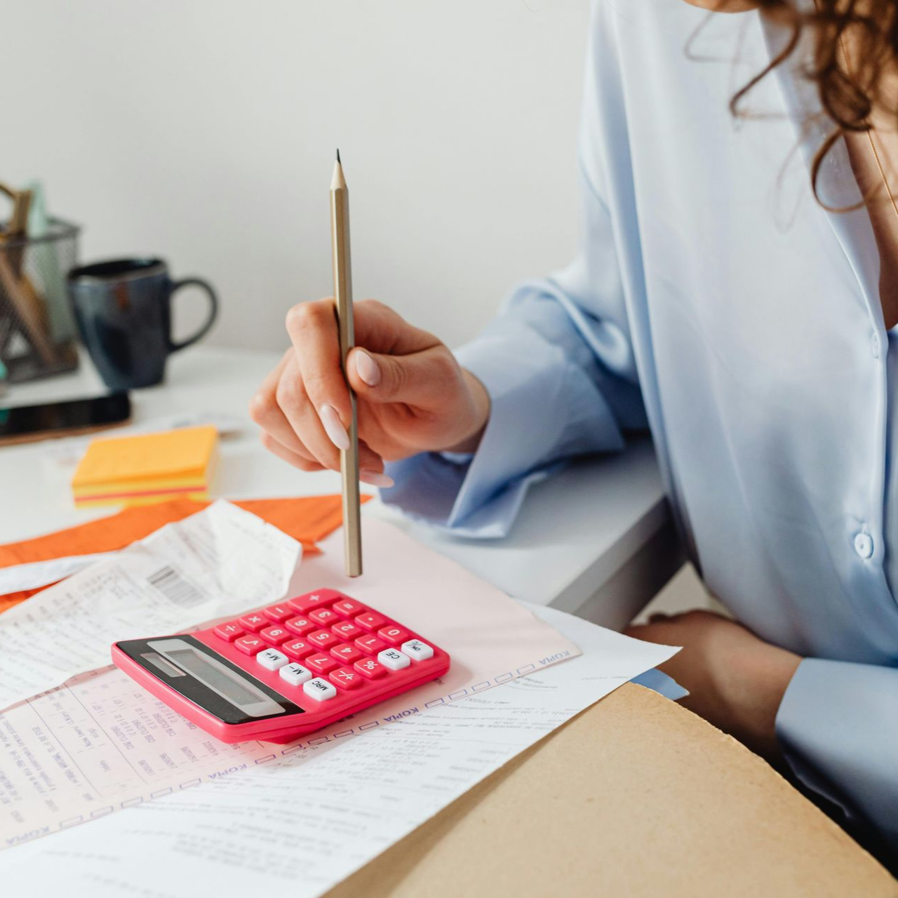 Woman in blue shirt using a pink calculator, with a pencil, papers, and coffee cup on a white desk.