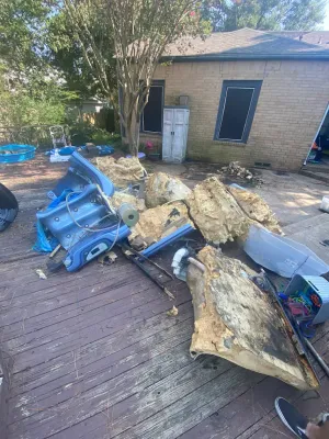 A pile of debris, including a blue plastic component and foam insulation, sits on a wooden deck outside a brick house.