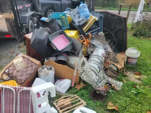 A large pile of assorted trash and discarded household items sits on the grass beside a parked truck.