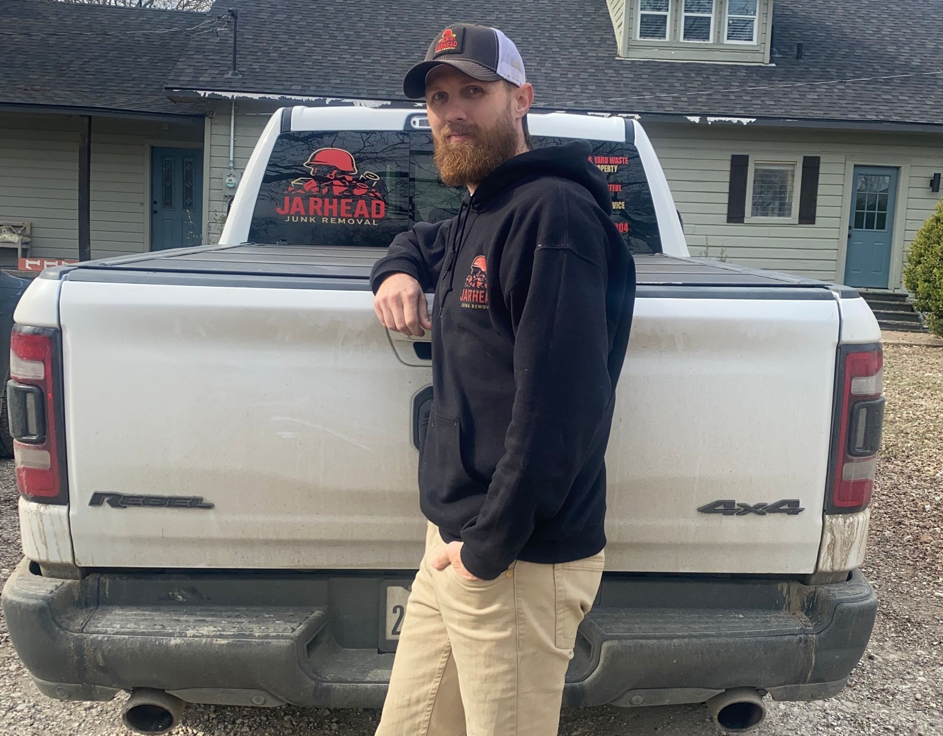 A bearded person in a hoodie and cap leans against the tailgate of a white truck parked in front of a house.