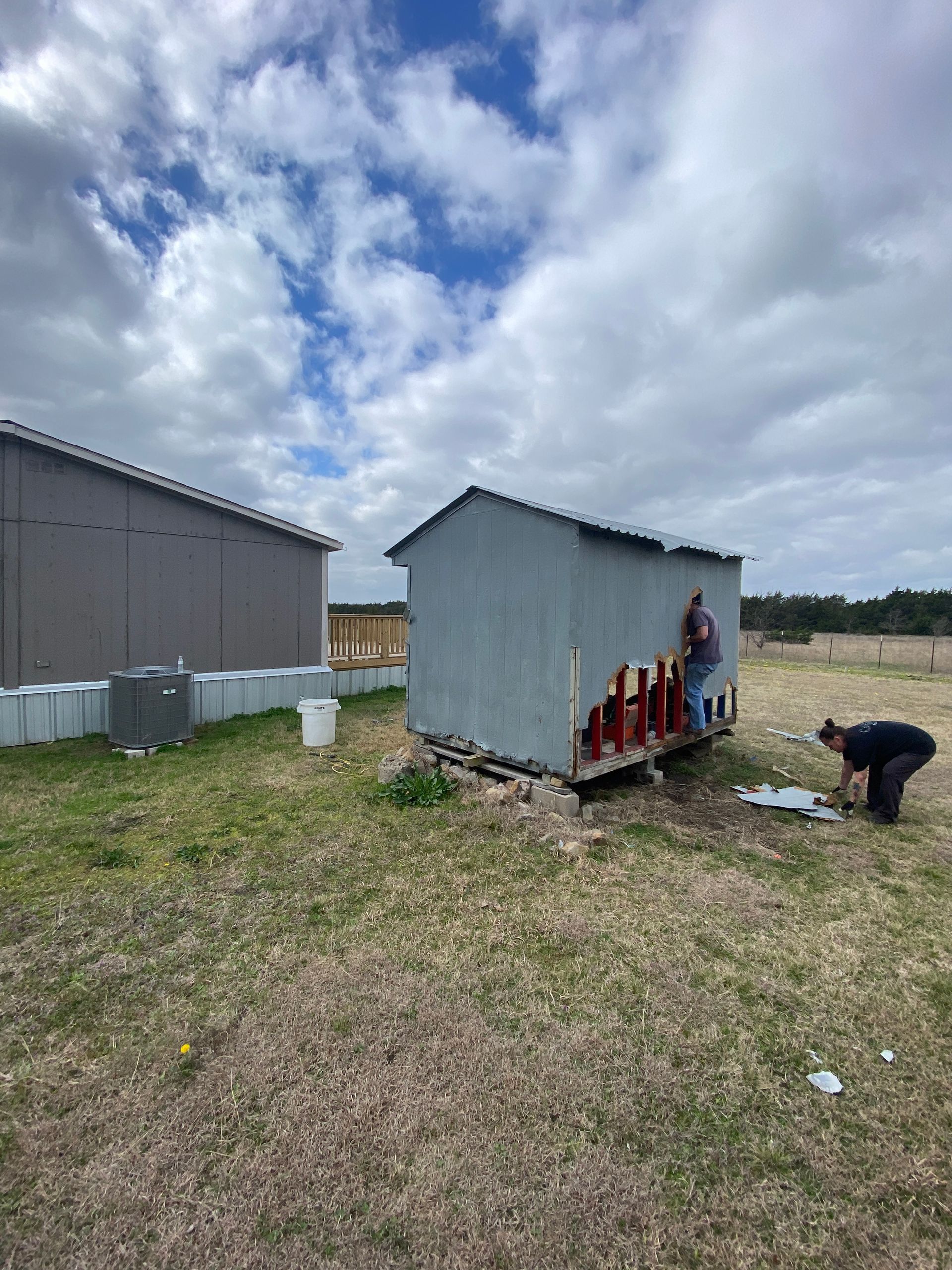 Two people work on repairing the siding of a small gray shed in a grassy field under a cloudy blue sky.