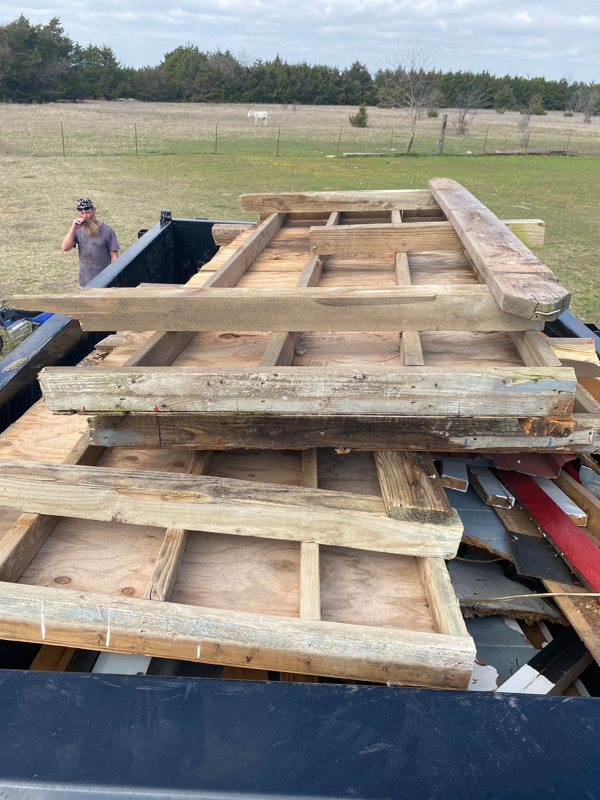 A person stands near a dump trailer filled with stacked, weathered wooden wall framing and plywood scraps in a field.