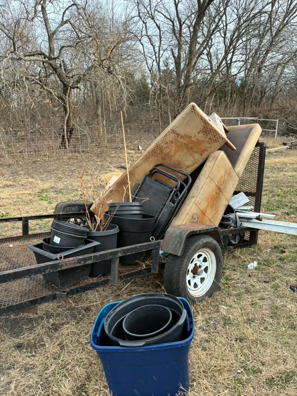 A utility trailer loaded with discarded household furniture and plastic pots, parked in an outdoor, grassy field.